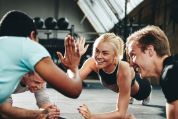 Eine Frau und vier Männer sind alle in der Plankposition und haben die Köpfe zueinander gewendet. Die Frau und der Mann gegenüber klatschen ihre Hände ab und sie lacht dabei. Sie befinden sich in einem modernen Fitnessstudio mit urbanem Charakter, einem grossen Dachfenster, weichem Sportbelag und vielen Fitnessgeräten im Hintergrund.  Eine Frau und vier Männer sind alle in der Plankposition und haben die Köpfe zueinander gewendet. Die Frau und der Mann gegenüber klatschen ihre Hände ab und sie lacht dabei. Sie befinden sich in einem modernen Fitnessstudio mit urbanem Charakter, einem grossen Dachfenster, weichem Sportbelag und vielen Fitnessgeräten im Hintergrund.